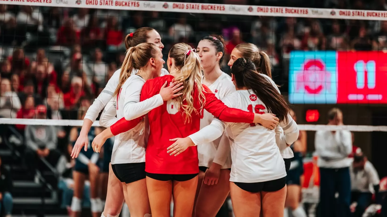 Ohio State volleyball players huddling at the net in front of the Ohio State Buckeyes banner during a Big Ten match