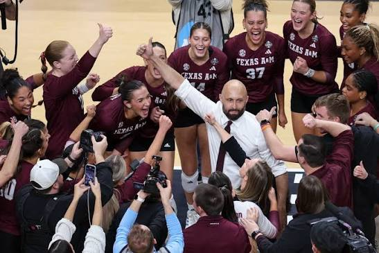 Texas A&M volleyball players and coaches on the sideline as the crowd stands behind them