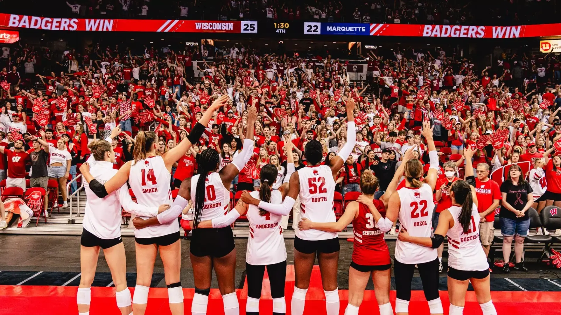 Wisconsin Badgers volleyball players raising their arms to a packed red-clad student section as the videoboard reads Badgers Win over Marquette