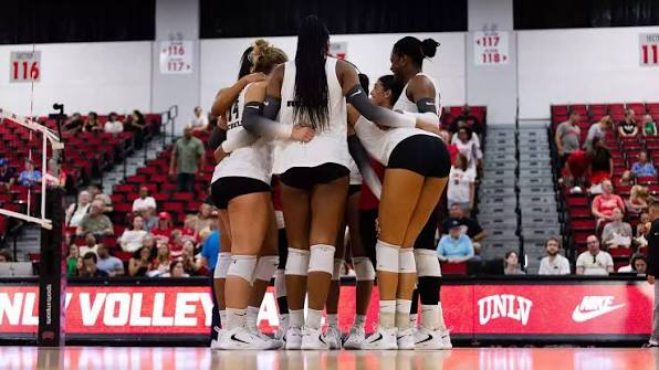 UNLV volleyball players huddling arm-in-arm in front of the UNLV Volleyball banner and red home crowd