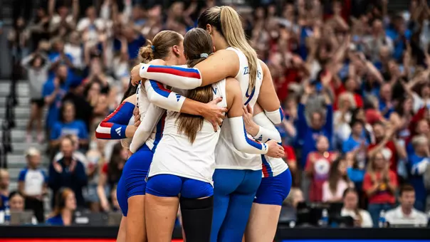 Kansas Jayhawks volleyball players huddling together in white and blue uniforms as fans cheer during a 2026 season match