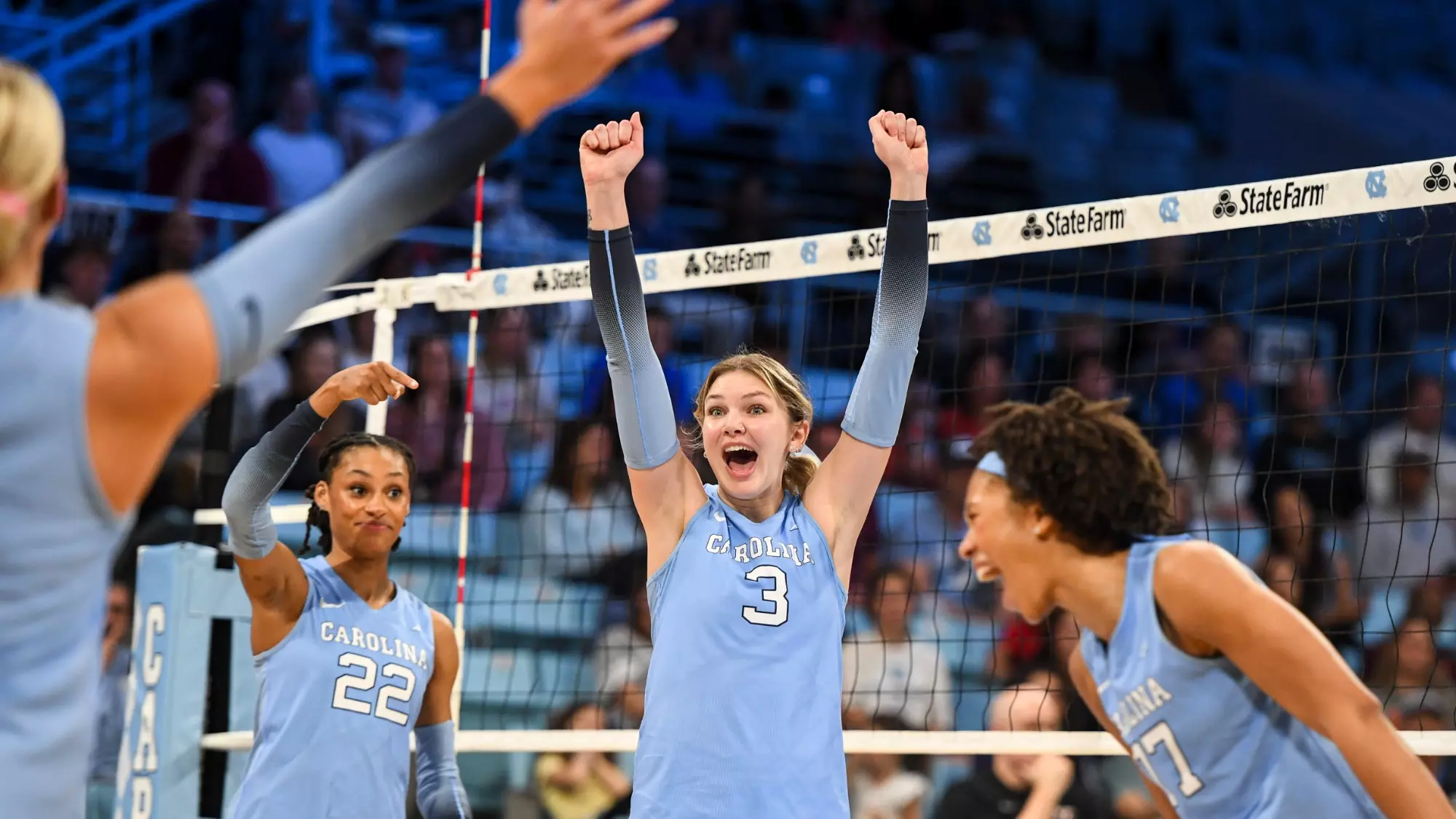 Carolina volleyball player No. 3 raising both fists in celebration with teammates at the net