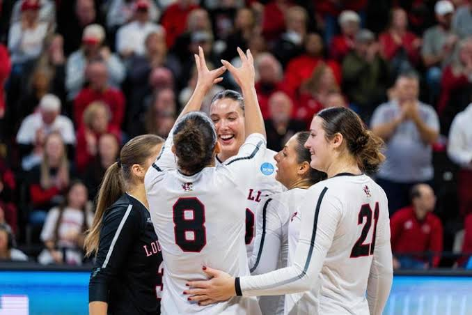 Louisville Cardinals volleyball team huddling after a rally win in front of a red-clad crowd