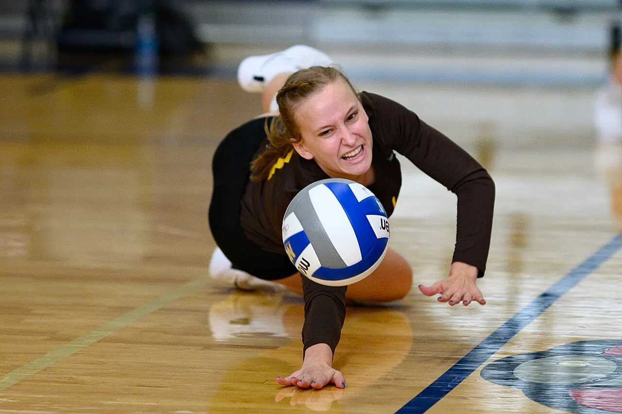 A Baldwin Wallace volleyball player reaching low with both arms to keep a ball off the court.