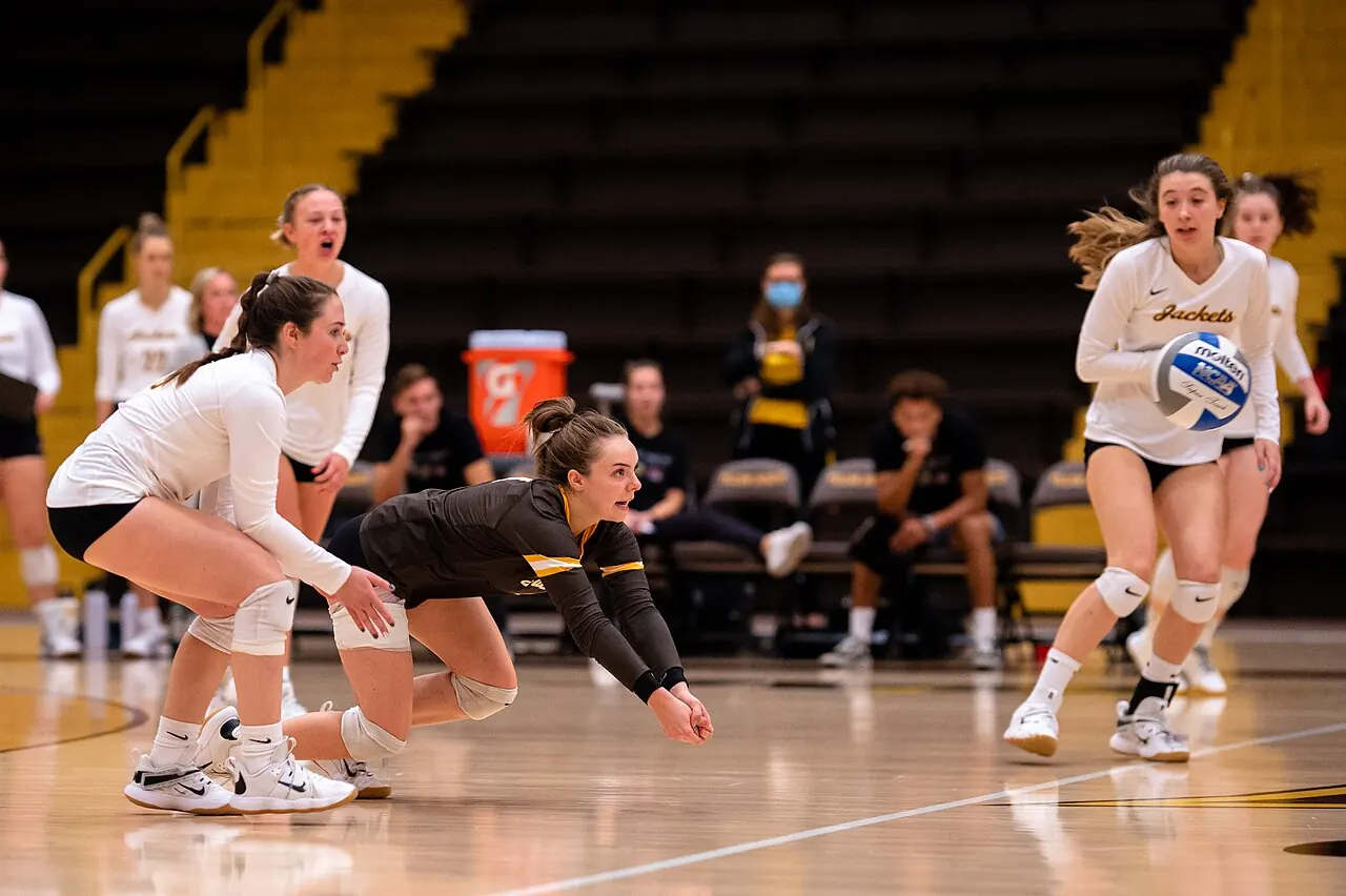 Baldwin Wallace volleyball players preparing to receive, with a black-jersey player behind white-jersey teammates.