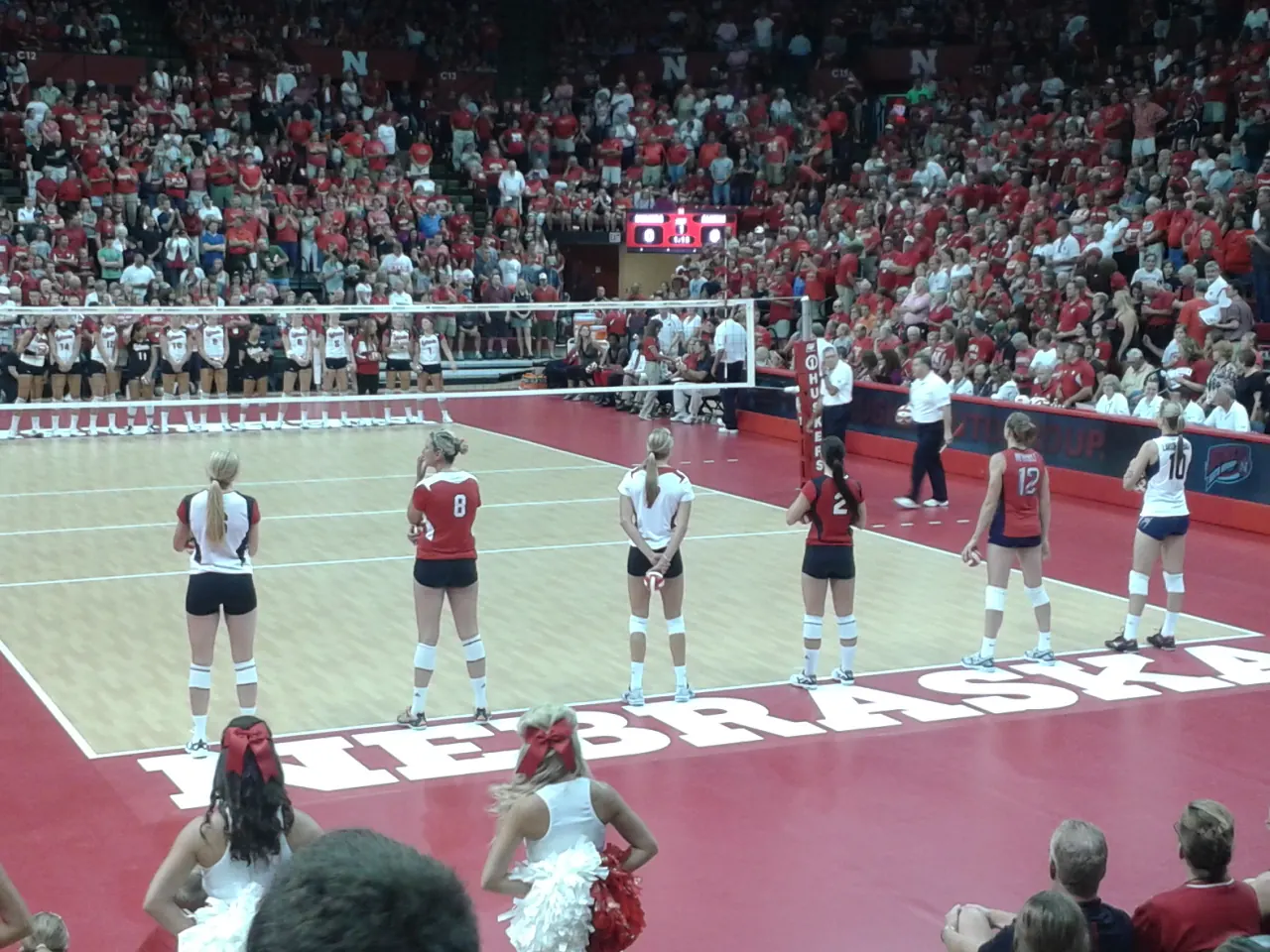 Nebraska volleyball players lined up beside the net at the Devaney Sports Center in front of spectators.