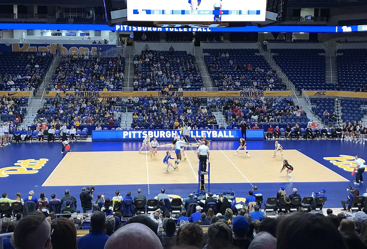 Pitt and Louisville volleyball players on court at the Petersen Events Center, with spectators in blue seats.