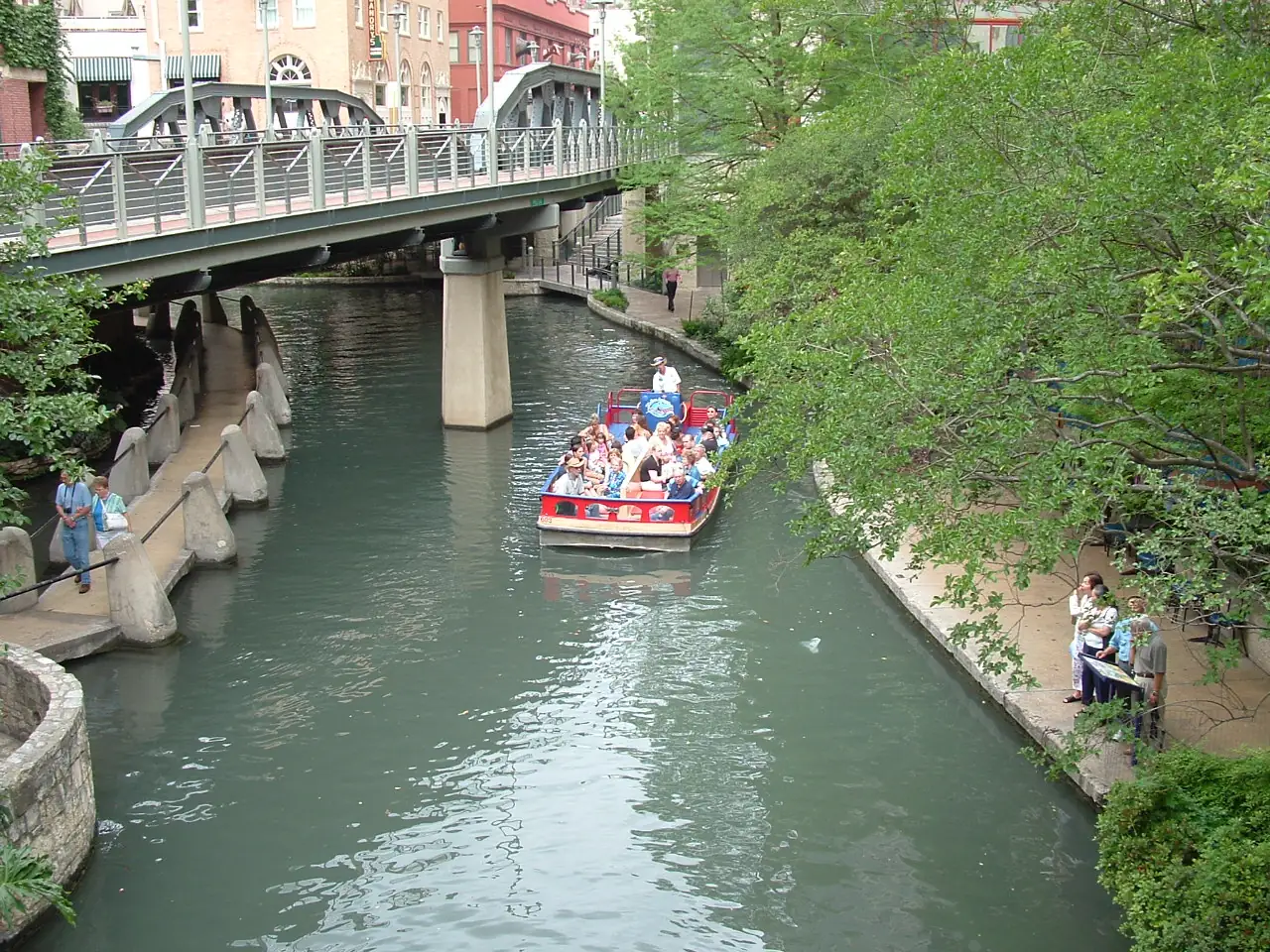 A sightseeing boat on the San Antonio River Walk beside trees and waterfront walkways.