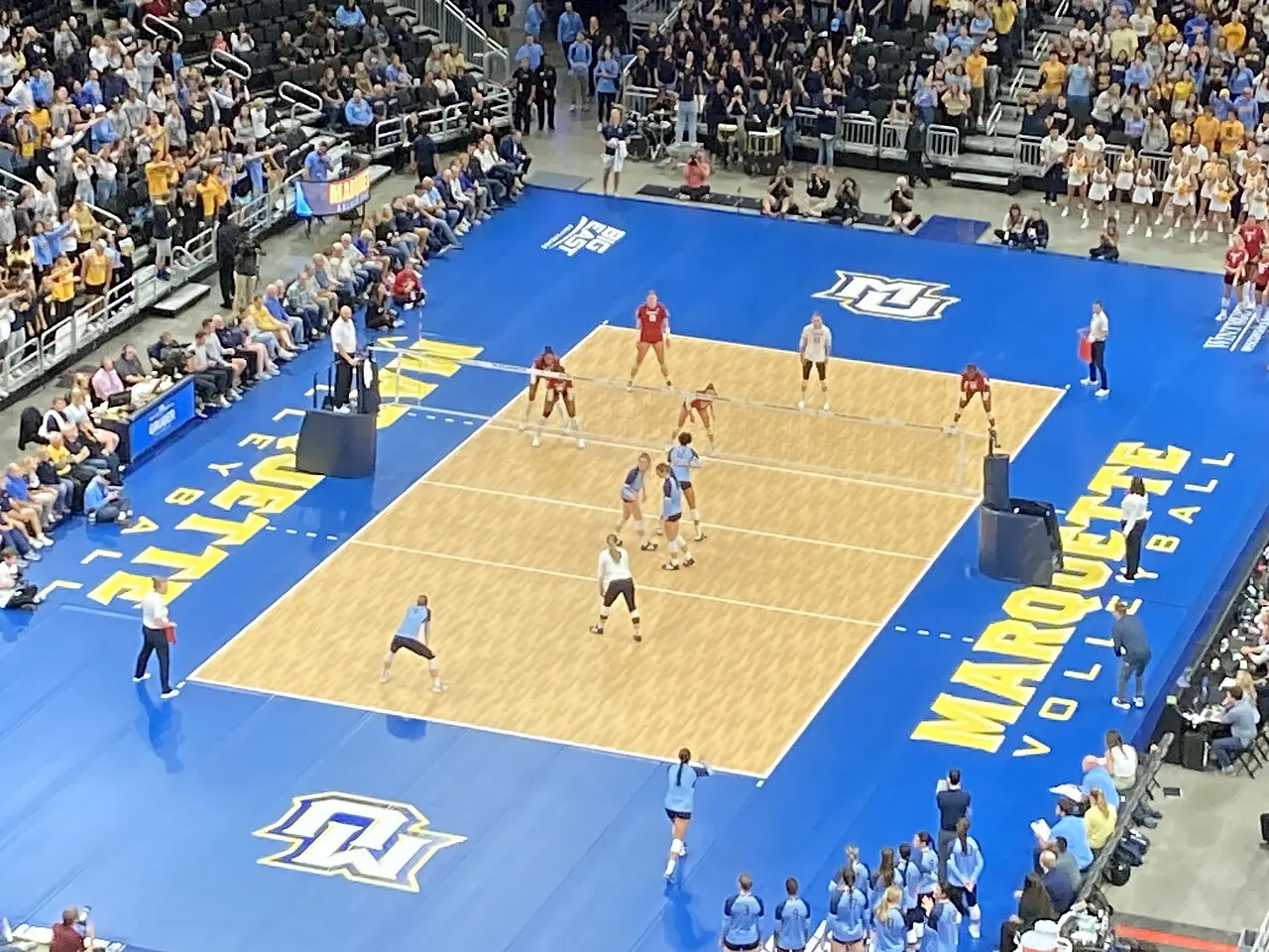 Wisconsin and Marquette teams arranged on opposite sides of the net with spectators surrounding the court.