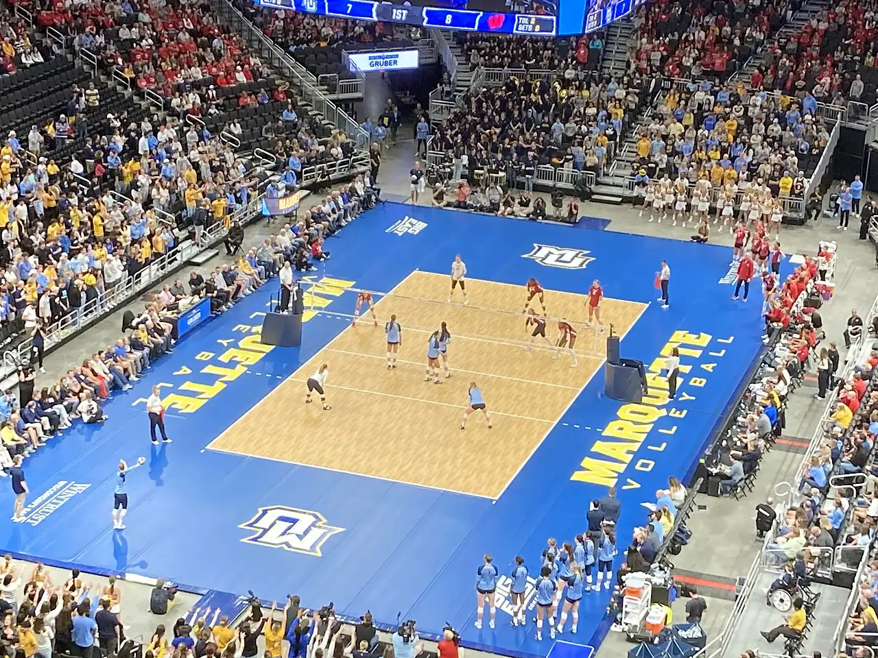 Wisconsin and Marquette players on a blue-and-gold volleyball court at Fiserv Forum.